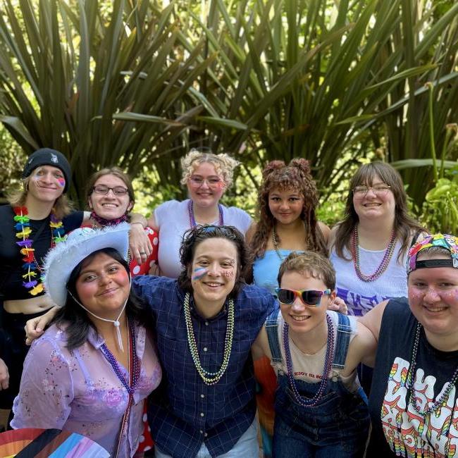 A group of students festooned in rainbow glitter and body paint smile at the camera. 